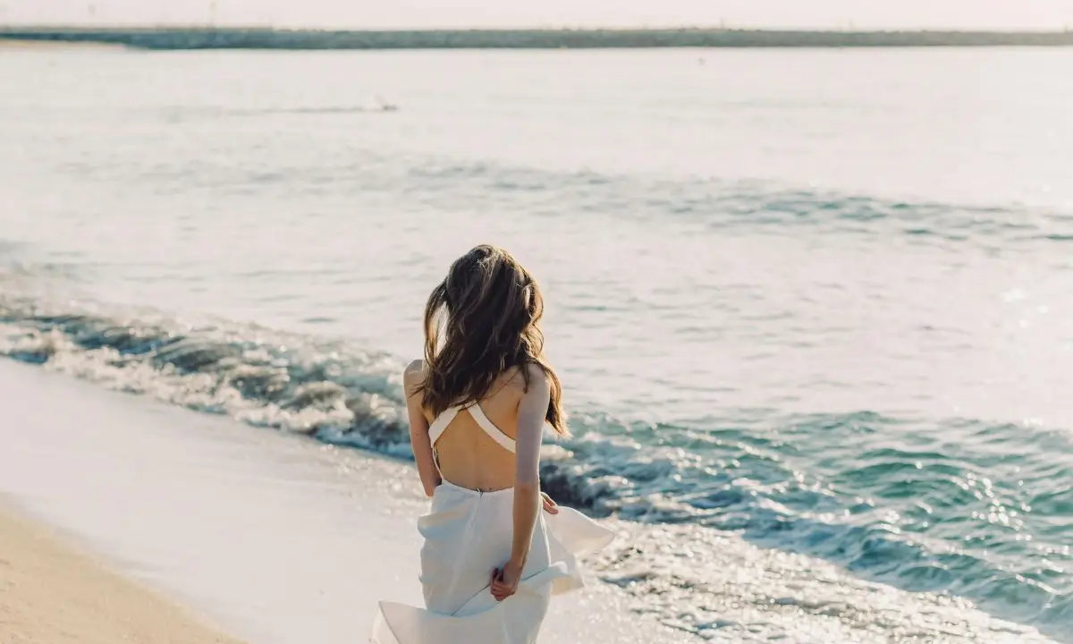 Donna con abito bianco che guarda il mare da una spiaggia luminosa